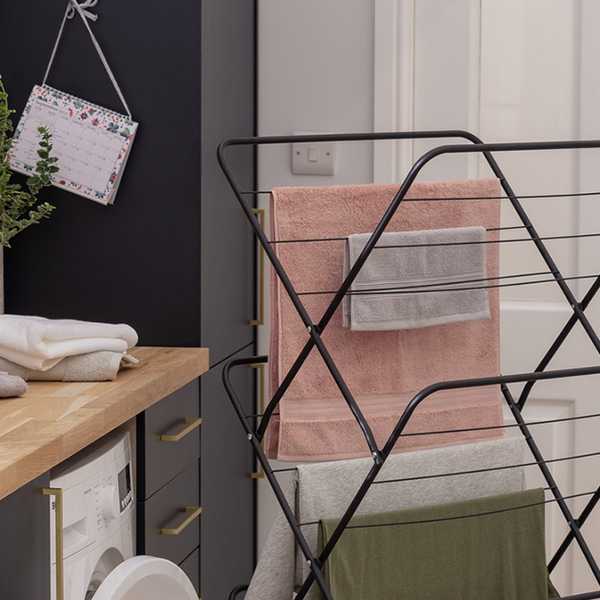An image of 3 tier indoor clothes airer in black with t-shirts and towels drying on it placed next to a washing machine. 